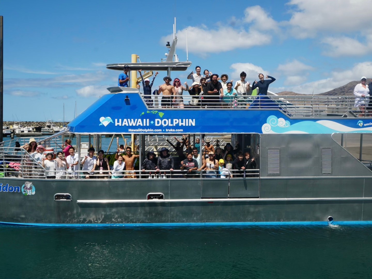 Tourists on a boat named Hawaii Dolphin in a marina with mountains in the background.