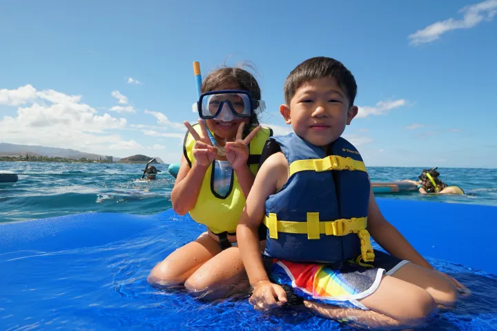 Two children wearing life vests sitting on a blue water mat in the ocean, with distant snorkelers.