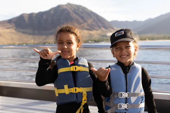 Two kids in life jackets smile and make shaka signs on a boat with mountains in the background.