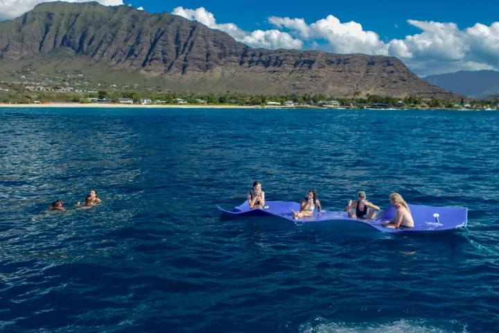 People on a floating mat and swimmers in ocean with mountains and shoreline in the background.