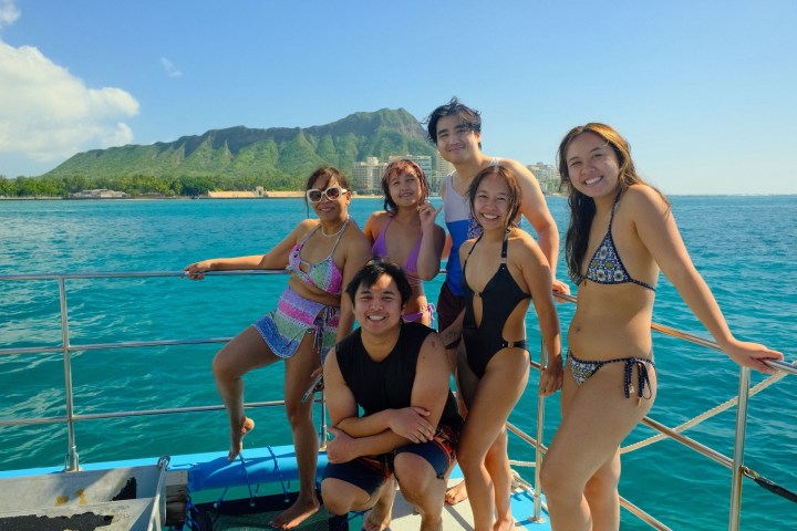 Six people in swimsuits on a boat, ocean and green mountains in the background.
