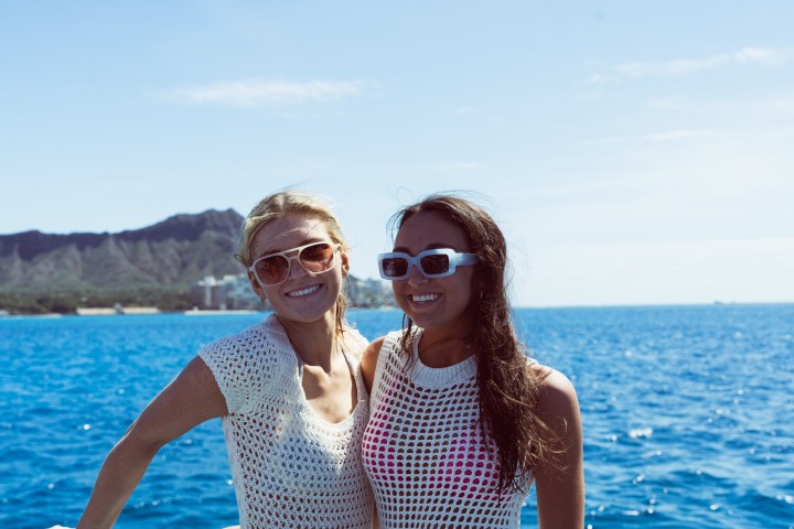 Two women in sunglasses smiling on a boat with ocean and mountain in background.