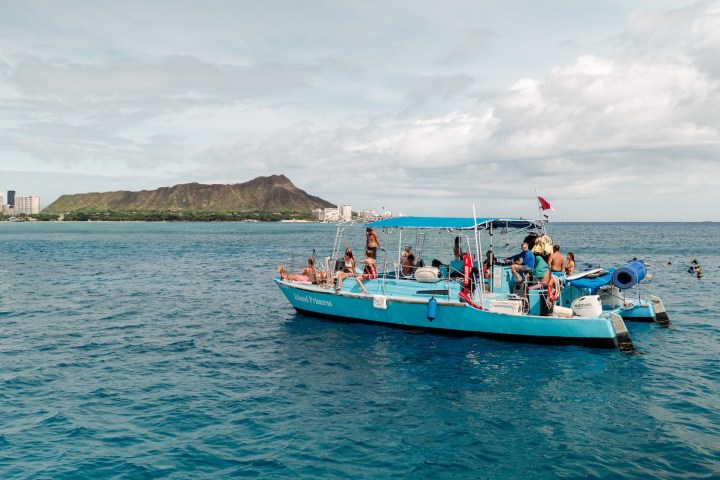 Boat with people on blue ocean near city skyline and mountain in background.