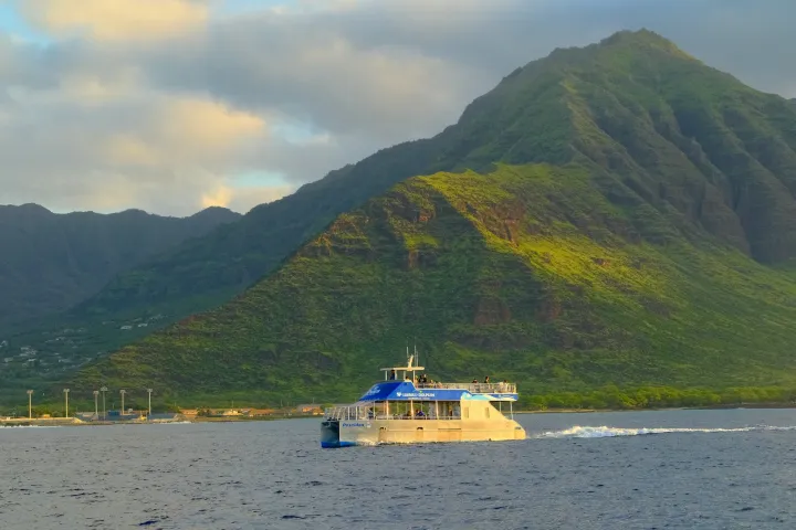 Boat on ocean with green mountain background under a cloudy sky.