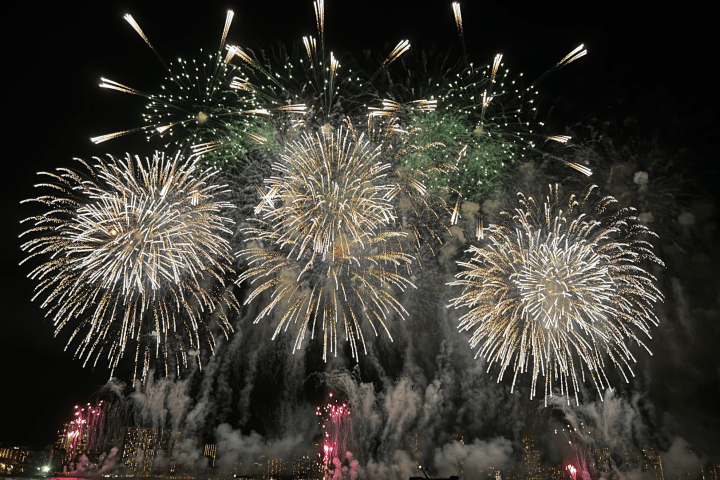 Fireworks exploding over a city skyline at night with boats on the water.
