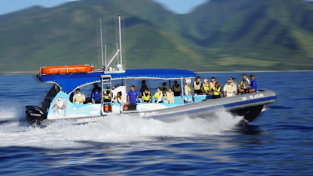 A speedboat with passengers wearing vests on blue water, with green hills in the background.