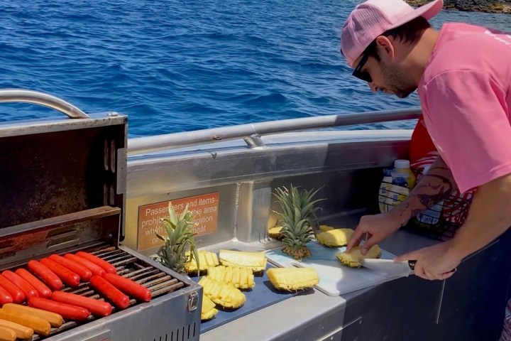 Person slicing pineapple on boat with grill cooking hotdogs, ocean in background.
