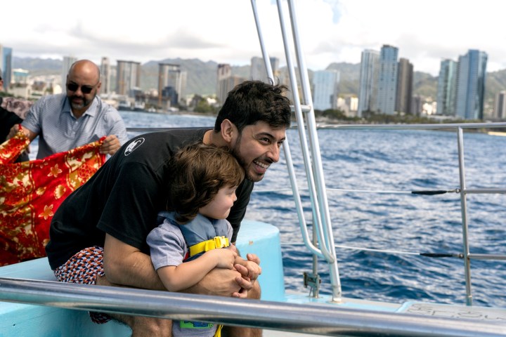 Man and child on a boat with city skyline in the background on a sunny day.
