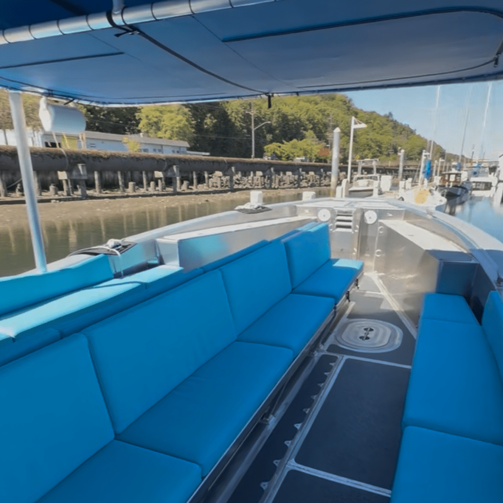 Interior of a boat with blue cushioned seating, docked by a waterfront with trees and other boats nearby.