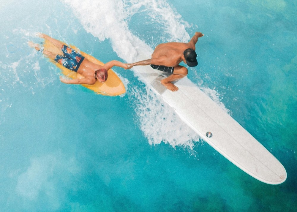 Two surfers, one on yellow board and other on white, join hands on ocean wave from aerial view.