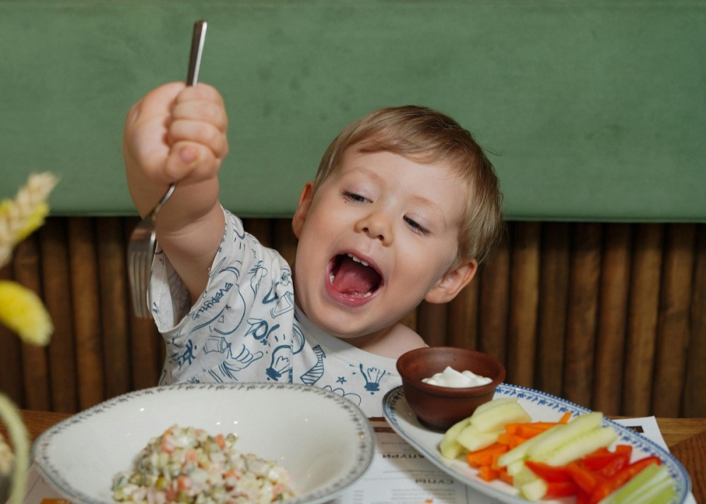 Child joyfully holds a fork over a bowl of salad with vegetables and dip on the table.