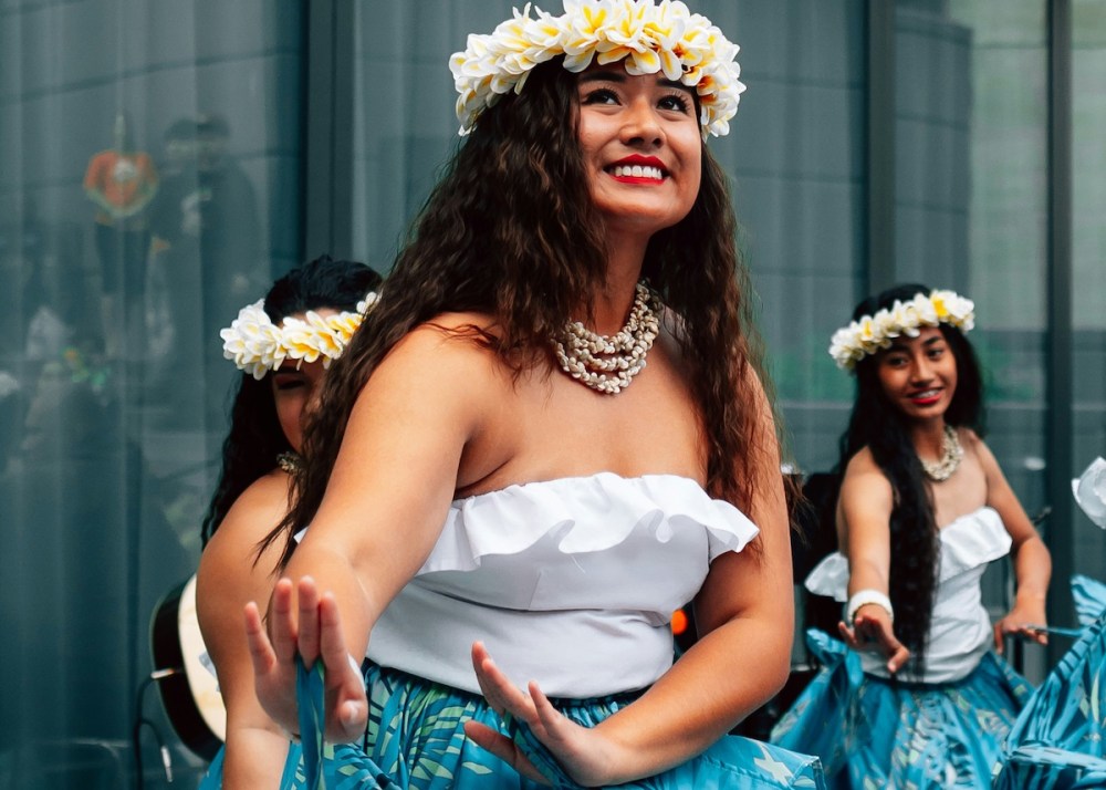 Women in blue skirts and floral crowns dance cheerfully in front of glass windows.