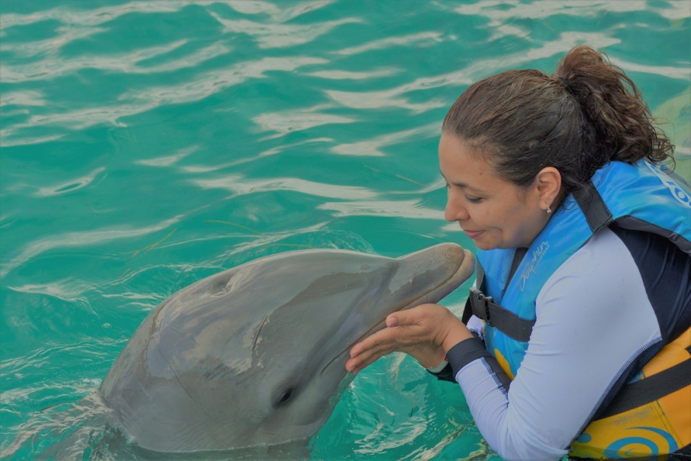 Woman in life vest gently touching a dolphin's face in turquoise water.