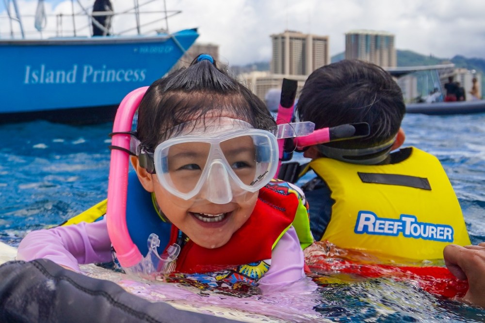 Two children snorkeling near a boat, wearing masks and floatation vests, smiling in the water.