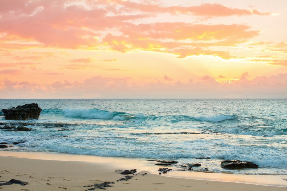 Sunset over ocean waves with rocks on a sandy beach.