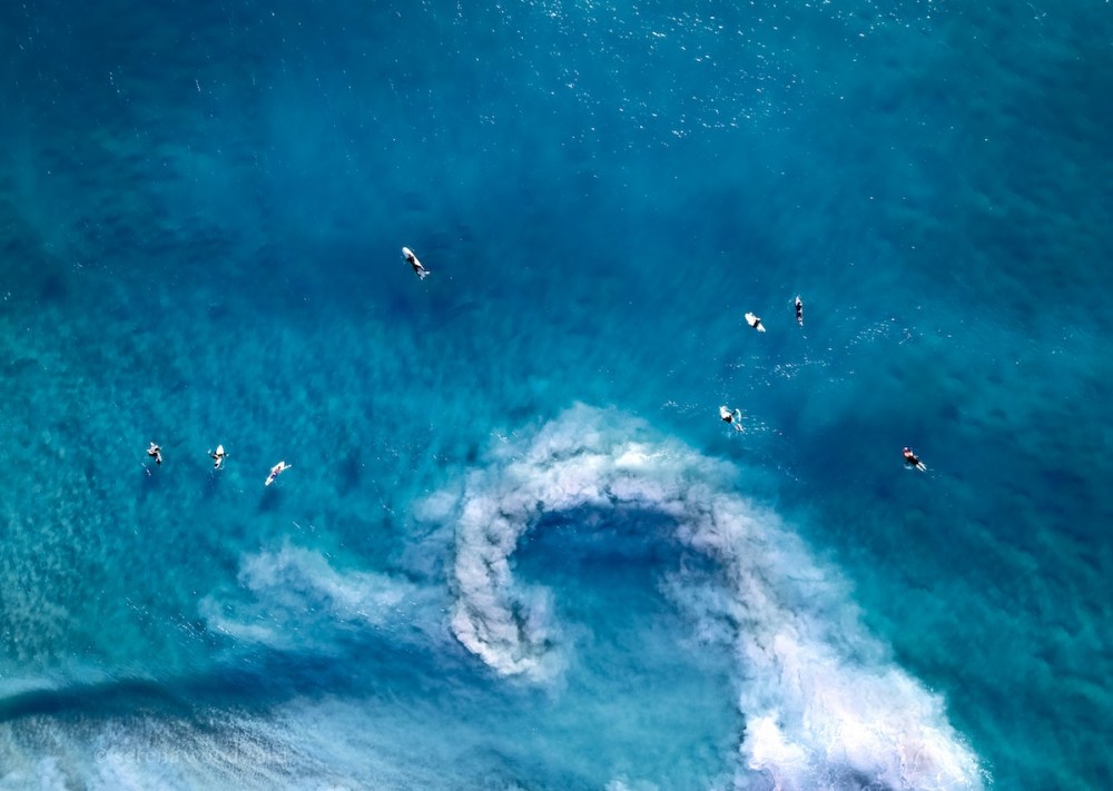 Aerial view of surfers paddling in blue ocean with a swirling wave pattern.